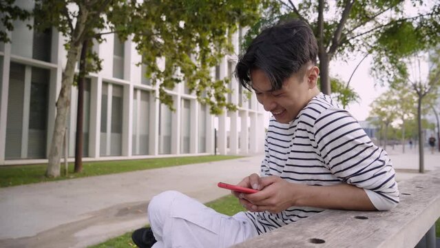 Funny Asian guy at university campus sitting on a bench chatting with a smart phone. Student using mobile cell and laughing outdoors. Cheerful people socializing on social media millennial generation