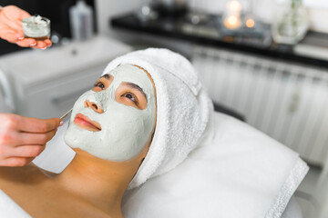 Deep purifying procedure at SPA. Portrait of beautiful positive smiling Asian woman in her 20s with white towel on her hair lying down on SPA bed while experienced cosmetician is putting clay mask on