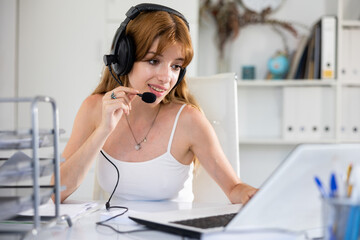 Naklejka premium Portrait of young woman wearing headset, using laptop. Call center worker sitting at desk in office.