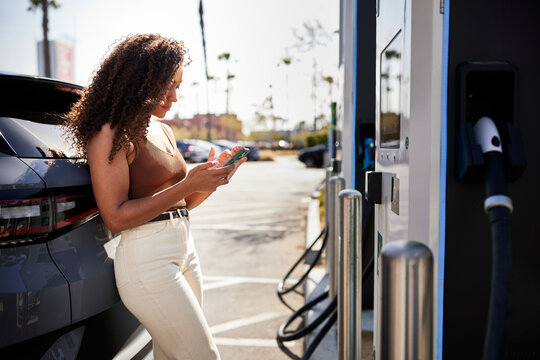 Young woman using phone leaning on electric car at charging station