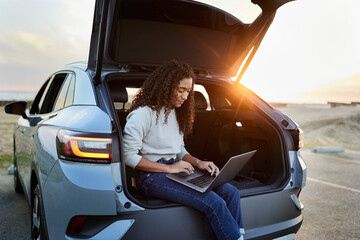 Young woman using laptop sitting in car trunk during sunset