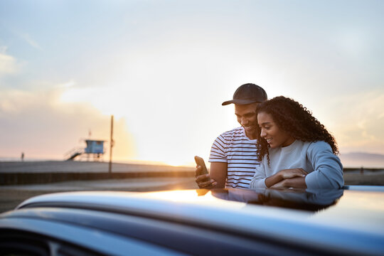 Smiling heterosexual couple using smart phone by car during sunset