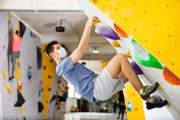 Male mountaineer in protective mask climbing artificial rock wall without his belay indoors