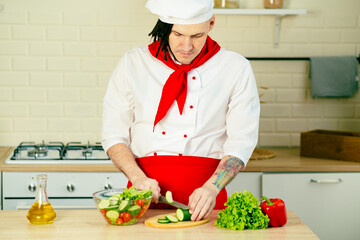 Young handsome chef cutting green cucumber on wooden board in kitchen. Positive guy with dreadlocks in uniform preparing vegetable salad.