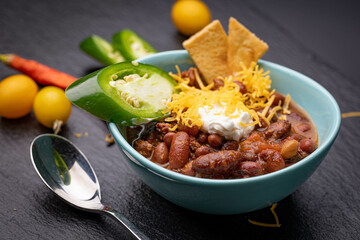 Small teal colored porcelain bowl filled with freshly cooked traditional homemade chili garnished with green jalapeño pepper, crackers, sour cream and shredded cheese