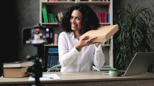 Pretty mixed race woman in casual wear recording video on camera while unpacking gift box. Female blogger sharing her emotions with her subscribers in social networks.