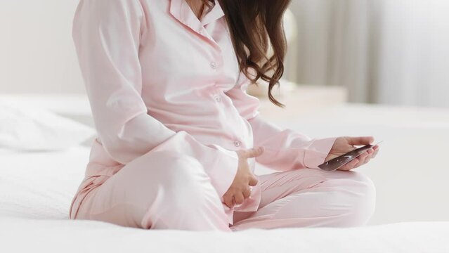 Unrecognizable pregnant woman in pajamas resting on bed, looking at sonography picture of her baby, empty space
