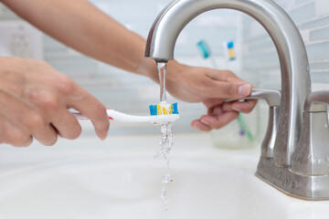 Hand holding a toothbrush while dipping toothpaste under the bathroom faucet.