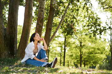 Relaxed young woman, resting near tree, sitting in park on lawn under shade, smiling and looking happy, walking outdoors on fresh air