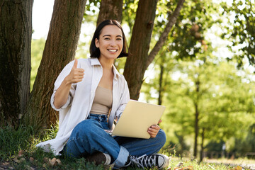 Portrait of asian young woman, student doing homework, working in park, sitting beside tree with laptop and showing thumb up, approve smth
