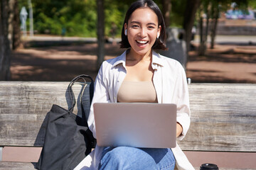 Portrait of asian woman sitting with laptop on bench in park, listening music with wireless headphones, doing homework, working on remote