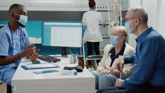 Nurse talking to woman with disability and her hurband ast consultation appointment during coronavirus pandemic. Specialist examining patient with impairment in wheelchair at checkup visit.