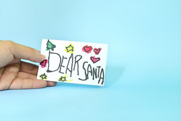 Christmas letter to Santa Claus from kid concept. Closeup of human hand holding a Dear Santa written letter drawing on white paper.