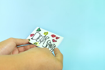 Christmas letter to Santa Claus from kid concept. Closeup of human hand holding a Dear Santa written letter drawing on white paper.