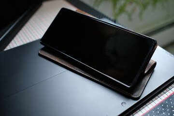 a cellphone in a brown leather case is stood up on top of a black lap top on a side table at home...