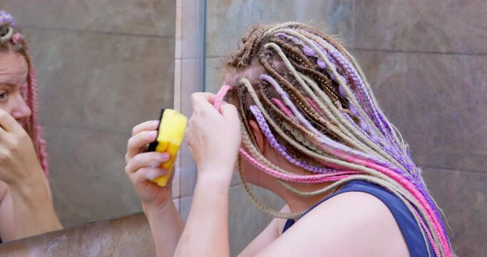 Woman with sponge in her hands stands in front of mirror, wipes her hair, washes her hair at home, care for afro braids. Cleansing and hygiene of hair with colored kanekalon. African braid care