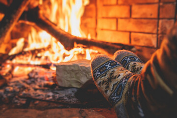 Warming up the fire, cozy winter night in the cabin house by the fireplace, fireplace burns in the scandinavian chalet cottage, burning fire with charcoal and firewood, view of woolen socks - obrazy, fototapety, plakaty