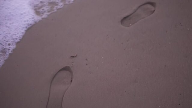 Slow-motion tracking shot of foot prints on the beach with waves crashing