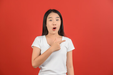 Portrait of Excited young asian woman in white t-shirt pointing finger at aside isolated on red background