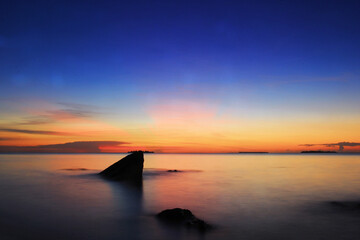 the view of the evening sky colored in gradations of dark blue and orange at the end of the beach with a rock silhouette