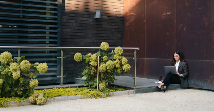 Far Away View Of A Young Woman Sitting Outdoor On Terrace, Working Remotely Online On Laptop. Freelance, Distant Job, Business Lady. Working From Home In Summer