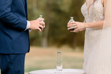 A bride and groom are holding two jars of sand right before they pour it to mix it for their...