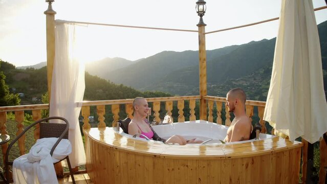 Young couple relaxing in jacuzzi on terrace with beautiful view of mountains. Attractive man and woman enjoying spending time together outside city during trip. Travel around the world