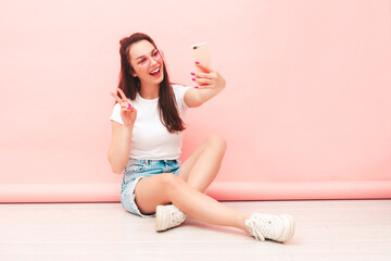 Obraz na płótnie Canvas Young beautiful smiling female in trendy summer hipster clothes. Sexy carefree woman sitting near pink wall in studio. Positive model having fun indoors. Cheerful and happy. Taking selfie photos