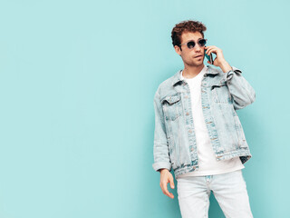 Handsome smiling stylish hipster model with curly hairstyle. Sexy man dressed in jeans jacket. Fashion male isolated on blue wall in studio. Holding smartphone. Talking at his phone