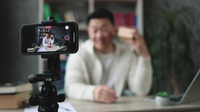 Focus on smart phone screen happy asian man in casual wear recording video on camera while unpacking gift box. Male blogger sharing his emotions with hs subscribers in social networks.