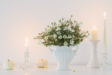 white flowers in white ceramic vase with burning candles in white interior