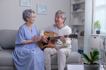 Asian mature male and female singing favorite song while playing on guitar at home. Elderly man and...