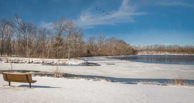 Panoramic view of Midwestern lake covered with melting ice on early spring day; wooden bench in foreground and blue sky with flying birds in background