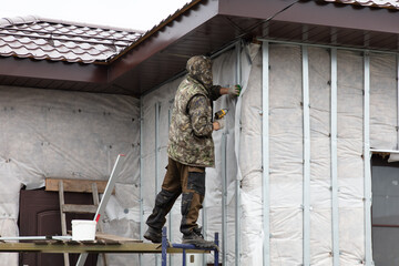 A man installs metal supports on the walls of the house for siding