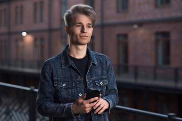 A young Caucasian blond guy in a denim jacket uses a mobile phone or smartphone.