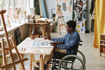 Obraz na płótnie Canvas Young contemporary artist in wheelchair working over new painting while sitting in studio of arts, mixing paints and applying them on canvas