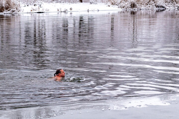 A middle-aged man bathes and swims in a winter river against the backdrop of snow-covered trees. The concept of hardening, healthy lifestyle. Bathing for baptism.