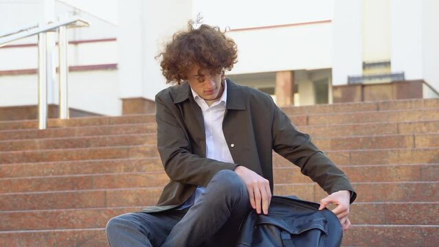 Young curly student puts books in a backpack near the university