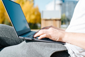 Man working on laptop outdoors. Close-up of male hands typing on keyboard while sitting on park bench, remote work. Side view, selective focus