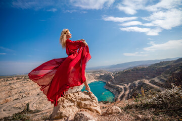 Fototapeta premium Side view of a beautiful sensual woman in a red long dress posing on a rock high above the lake in the afternoon. Against the background of the blue sky and the lake in the form of a heart