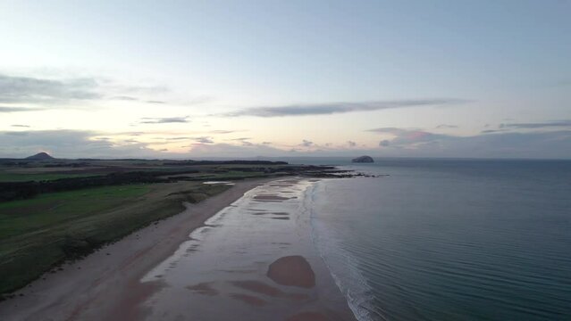 High panning drone footage flying above a long sandy beach at sunset slowly turning to look out across the rippling ocean as the tide gently laps the shore. Tyninghame Beach, Scotland