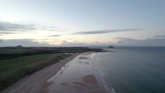 Stationary drone footage flying high above a long sandy beach at sunset looking out across the rippling ocean as the tide gently laps the shore. Tyninghame Beach, Scotland