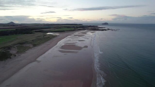 Fast aerial drone footage flying high above a long sandy beach at sunset looking out across the rippling ocean as the tide gently laps the shore. Tyninghame Beach, near Dunbar, Scotland