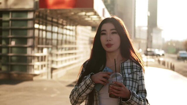 Young successful asian girl walks along the city street holding a glass with a refreshing drink in her hands. Confident girl. Career people. High quality 4k footage