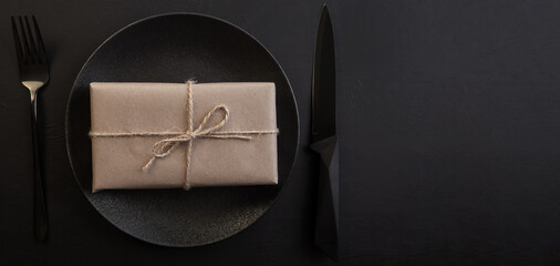 Gift box in a plate with a fork and knife on a black background. Creative table setting. Holiday concept. Place for text or advertising. Christmas. Valentine's Day.