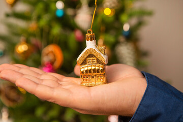 A golden holiday house held by a hand on the background of a Christmas tree. - 546200461