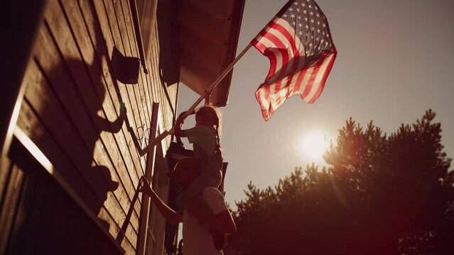 Proud Patriotic Dad Holding His Young Daughter of His Shoulders, Helping Her to Put the United States of America Flag on the Wall of Their House to Celebrate a National Holiday. Sunny Day in USA.