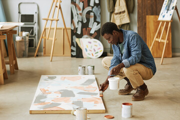 Young black man sitting on squats and bending over canvas with unfinished artwork while painting it in studio or classroom - obrazy, fototapety, plakaty