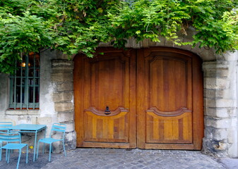Porte cochère en bois clair surmontée de verdure avec table et chaises bleues en fer.