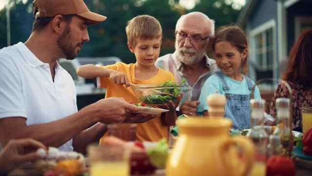 Young Kid Picking Up a Bowl of a Tasty Vegan Salad. Son Putting Vegetarian Food on a Plate to His Father. Outdoors Dinner Party with Family Members, Relatives and Multiethnic Friends.